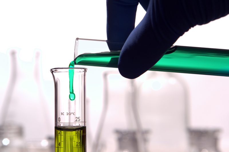 Scientist hand pouring a green chemical solution from a laboratory glass test tube into a scientific cylinder with yellow liquid for an experiment in a science research lab